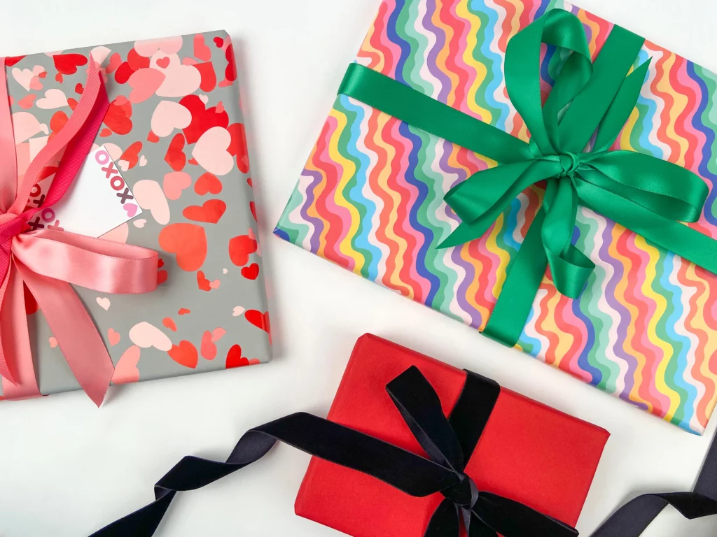 Emily in Paris inspired gift wrapping looks. pictured are three gifts, one wrapped in confetti hearts gloss wrapping paper styled with lipstick pink satin ribbon and fuschia pink grosgrain ribbon with an XO gift tag, another gift wrapped in squiggly rainbows gloss wrapping paper styled with emerald green satin ribbon, and the last gift is wrapped in printed kraft wrapping paper plain red styled with woven edge velvet ribbon black. pictured on a white background.